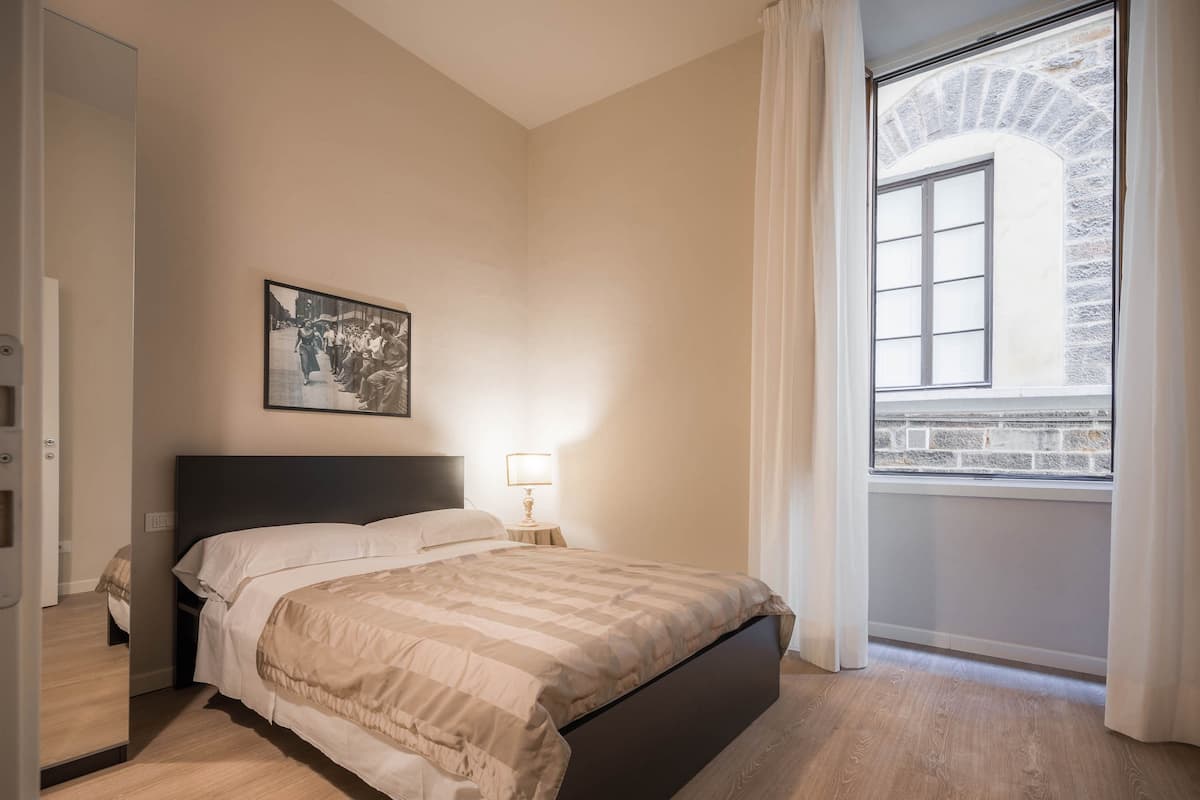 A bedroom with a bed, a nightstand, a lamp, a window with curtains, and a picture on the wall.