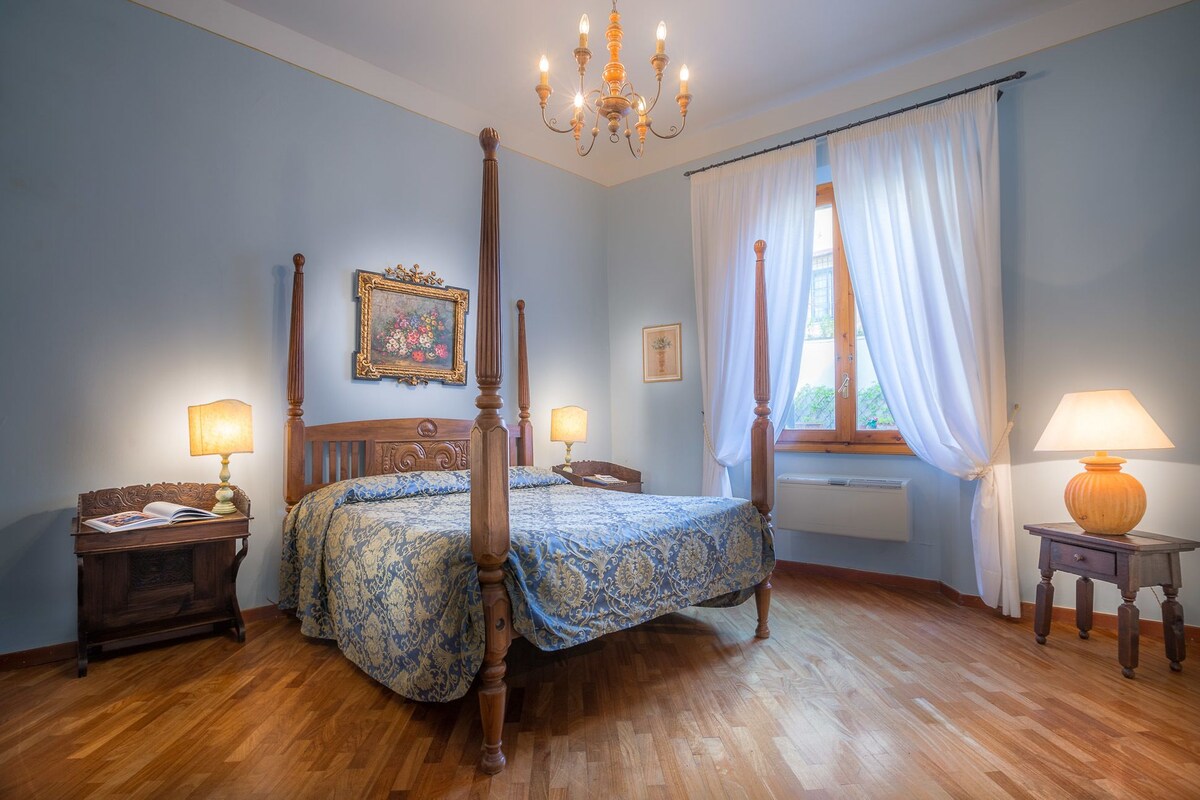 A four-poster bed with a floral bedspread, two bedside tables with lamps, a window with curtains, and a chandelier.