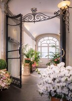 An open wrought iron gate leading to a sunlit interior with potted plants and a large arched window.