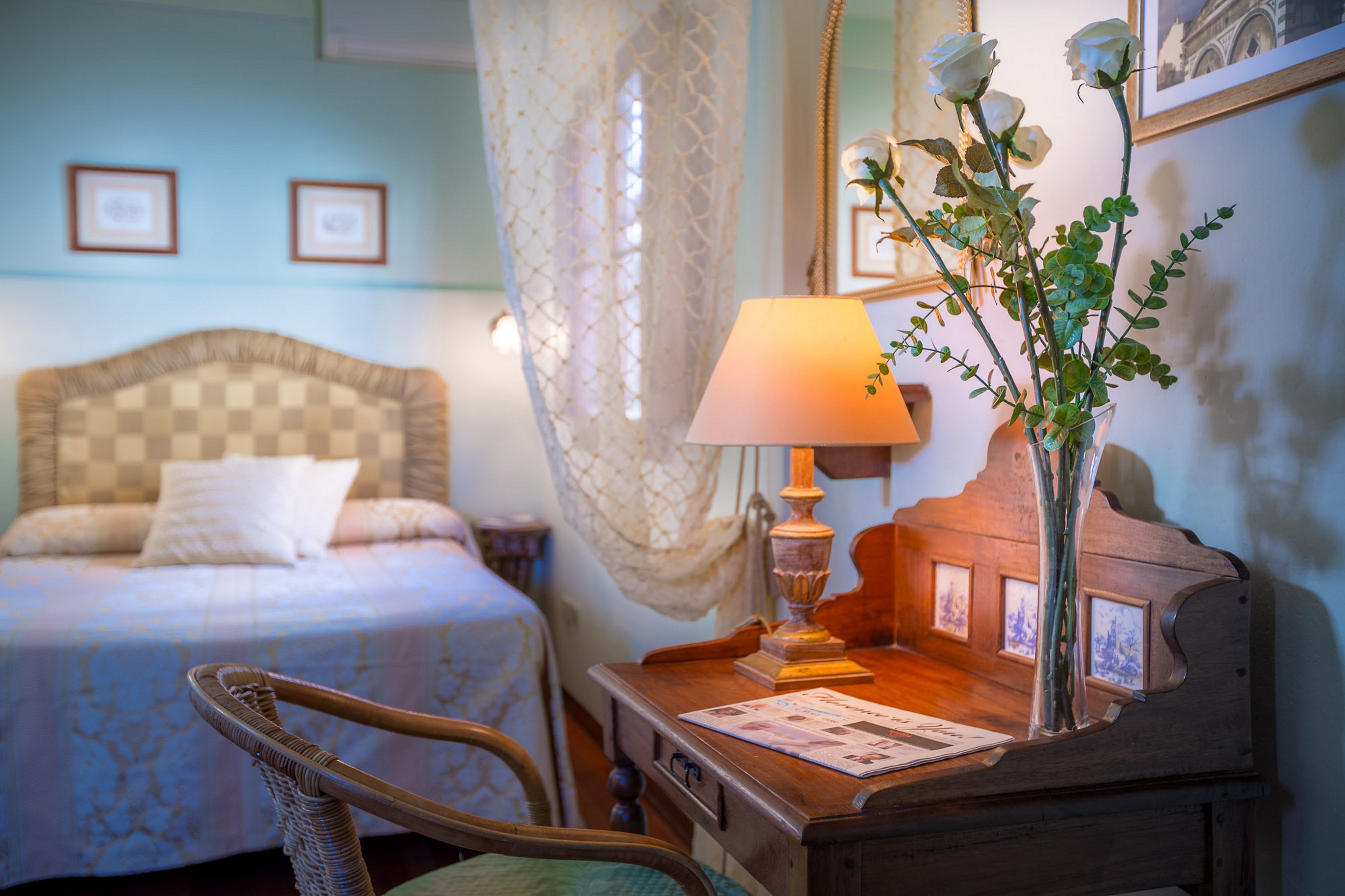 A bedroom with a bed, a wooden nightstand, a lamp, a vase with flowers, and a chair.