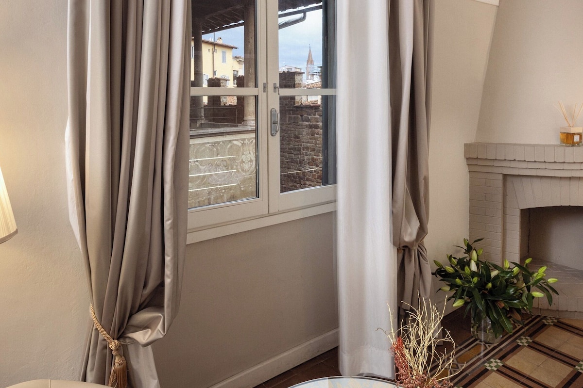 A room with a window, curtains, a fireplace, a table with a tray, a chair, and a vase with branches.