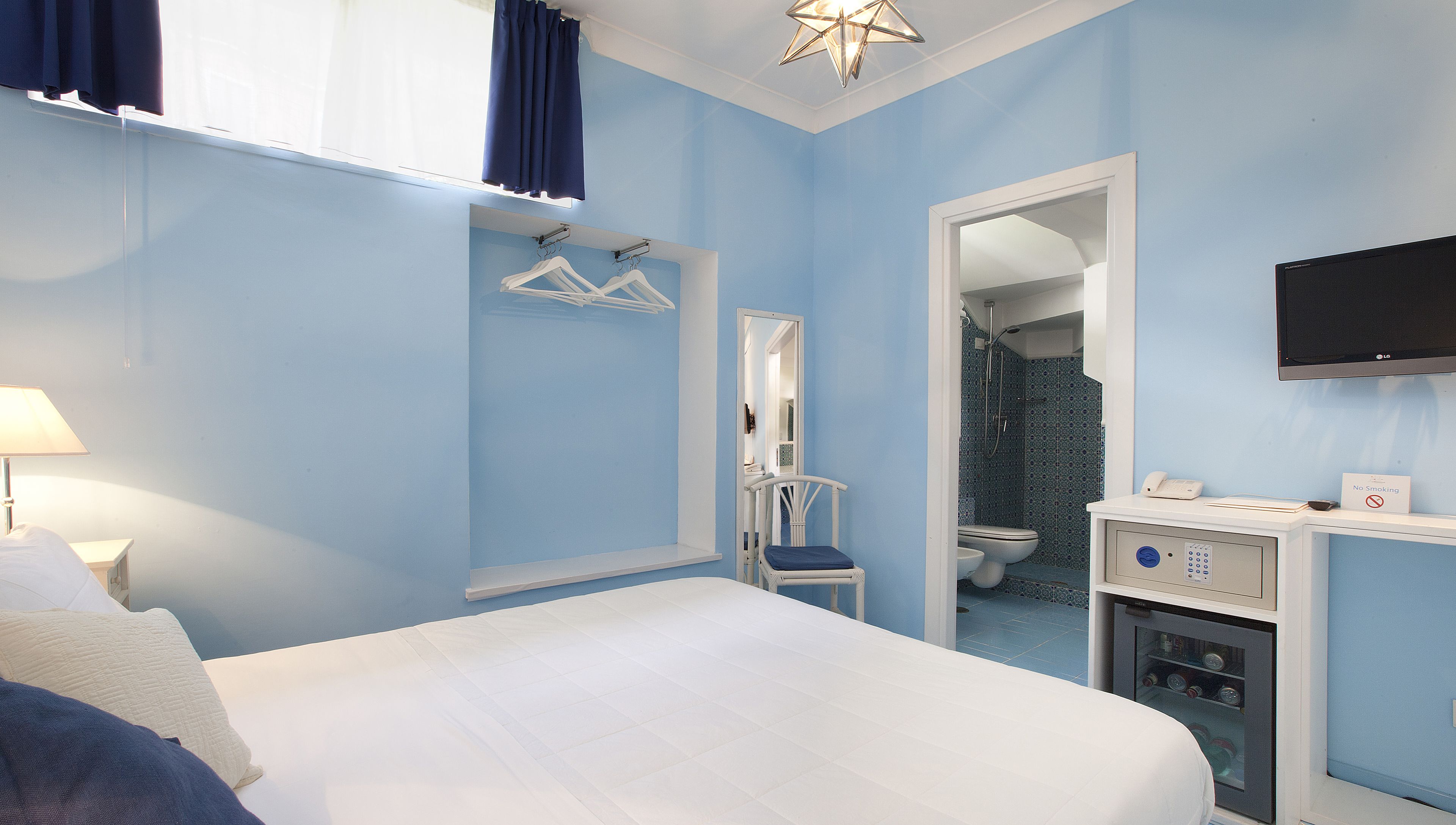 Habitación de hotel con cama, una silla, escritorio con teléfono, televisión y baño visible a través de una puerta abierta.