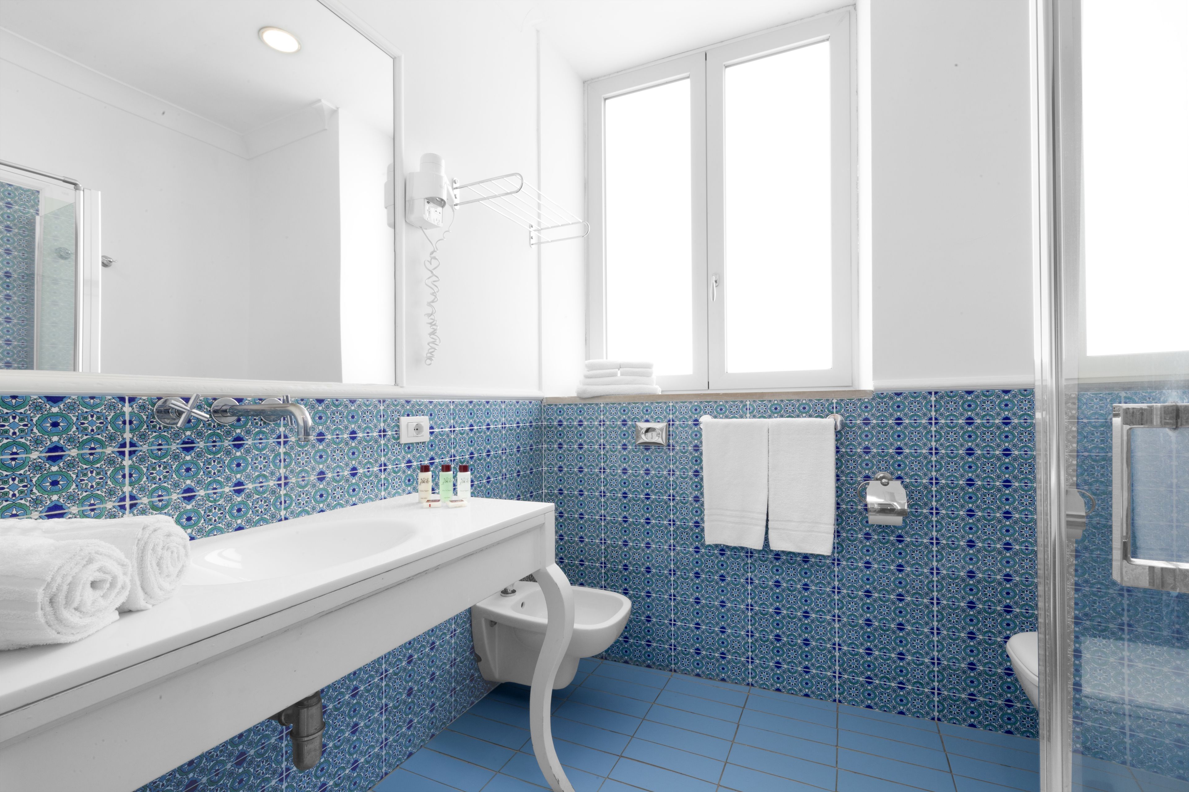 A bathroom with a white sink, blue-tiled walls, a glass shower enclosure, and a window.