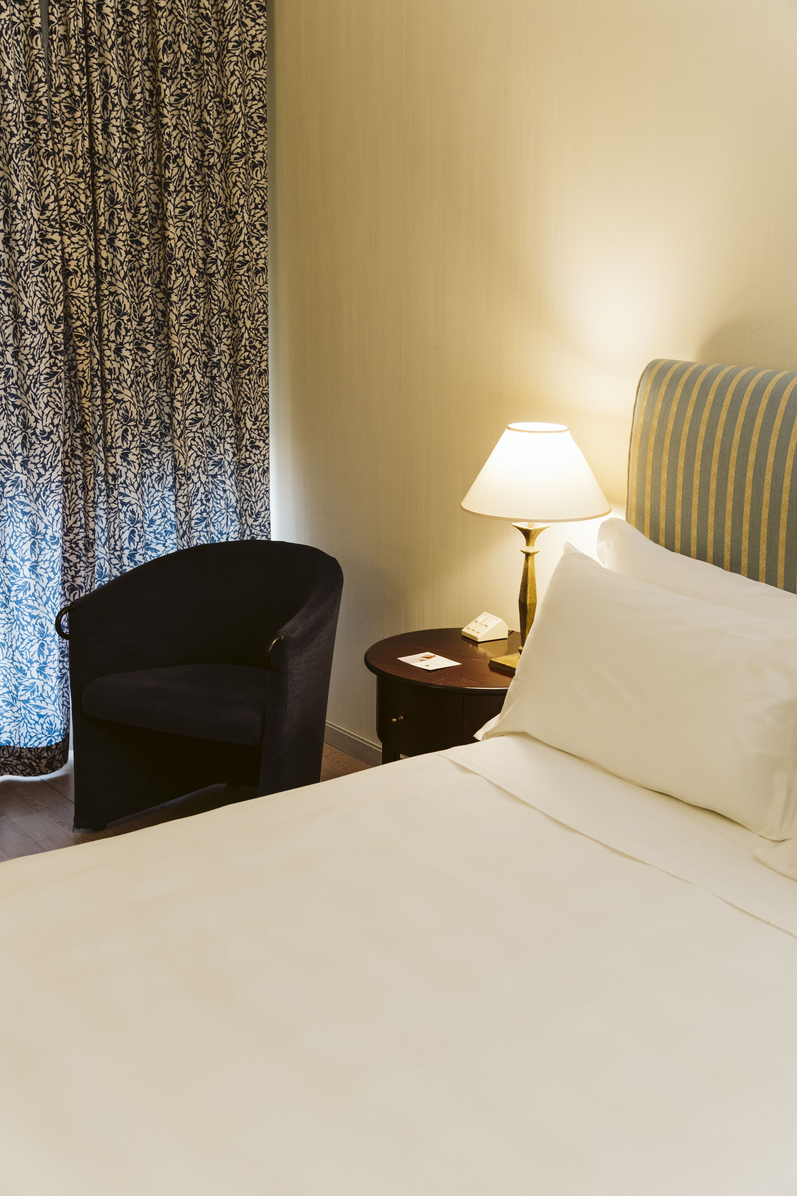 Ein Hotelzimmer mit einem Bett, einem Sessel, einem Nachttisch mit Lampe und einem gemusterten Vorhang.