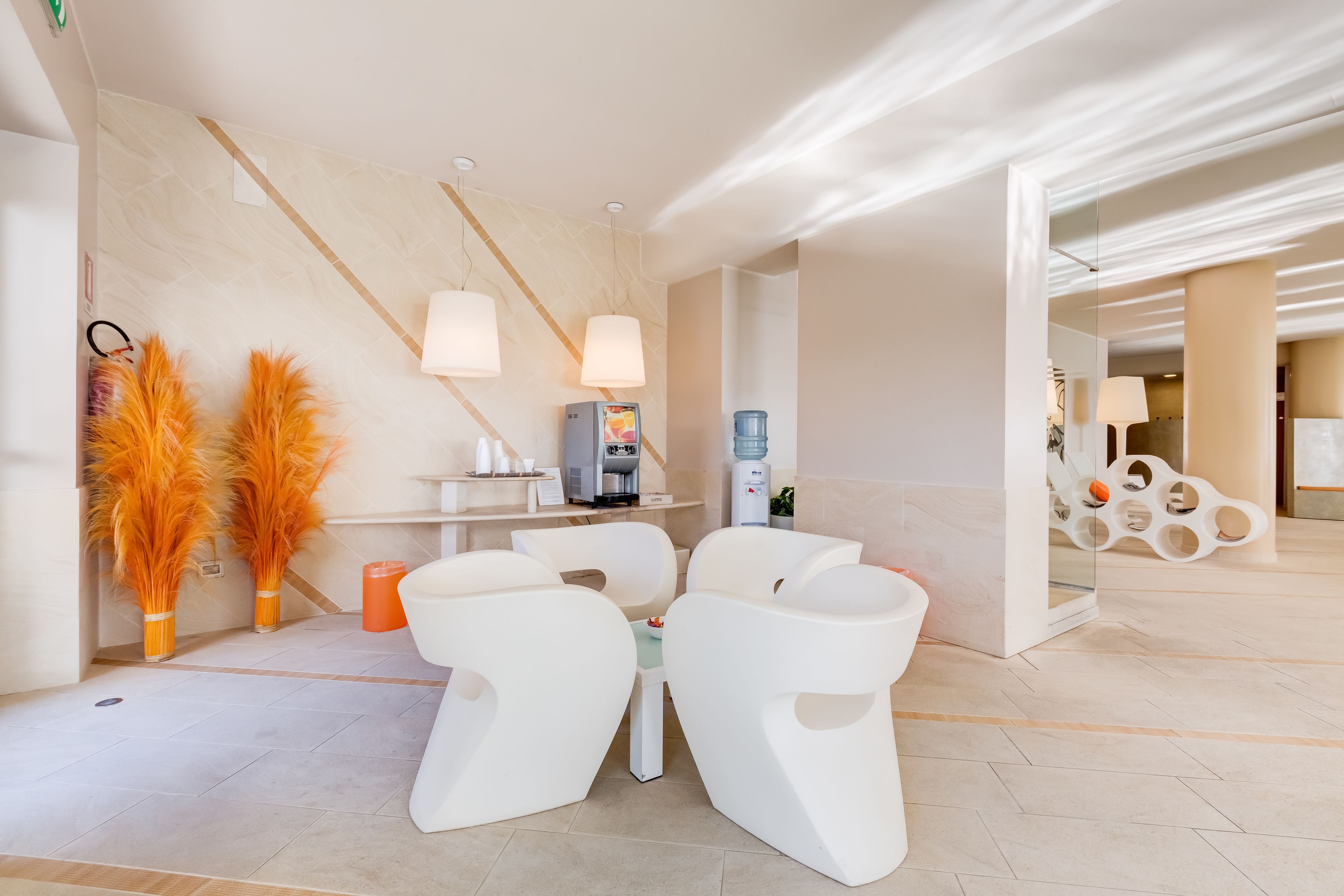 Una hall d'albergo moderna con una reception, una macchina per caffè e un erogatore d'acqua. Due sedie bianche e un tavolino si trovano al centro.