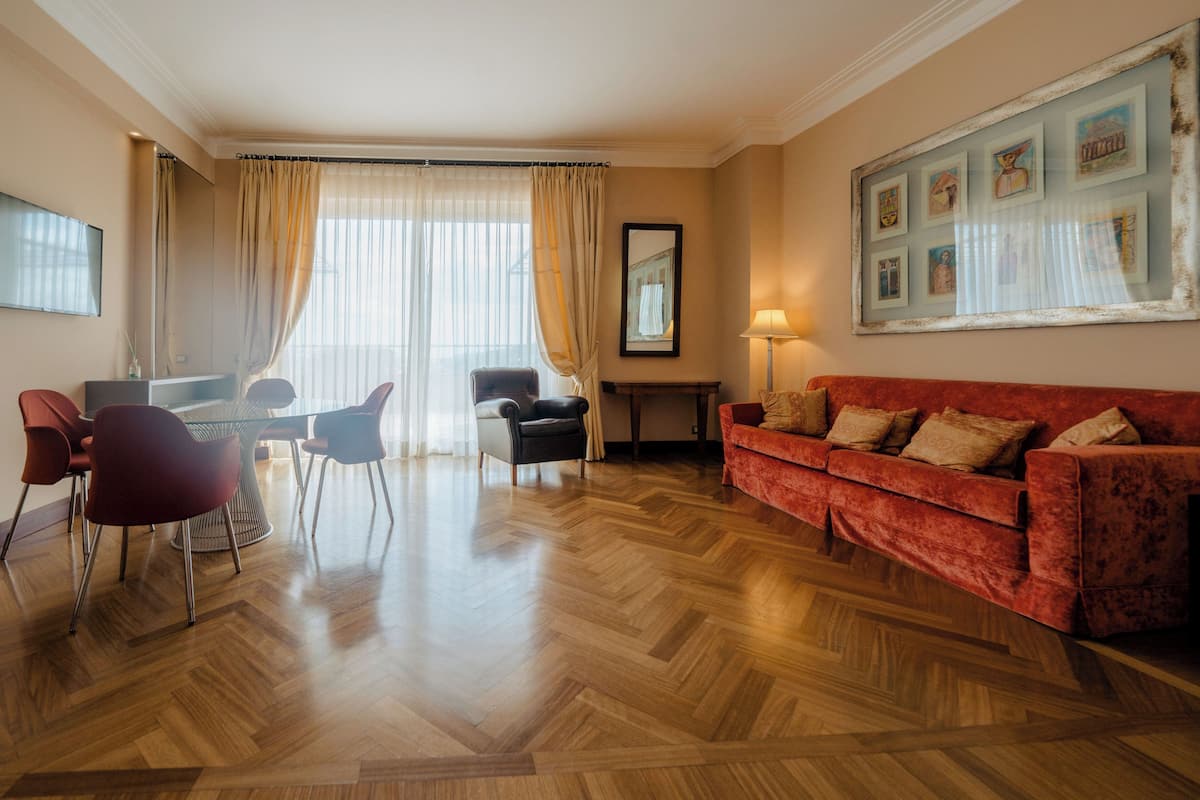 A spacious living room with a wooden parquet floor, a red sofa, a dining table with chairs, and a large window with curtains.