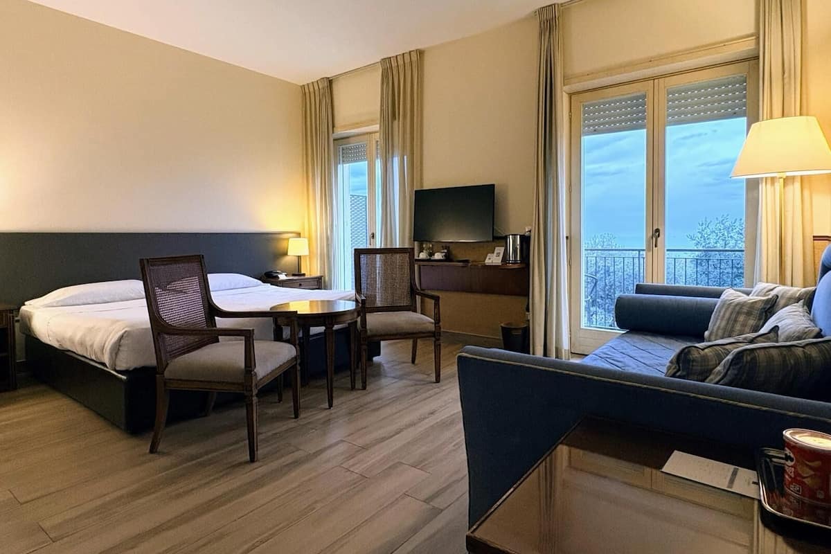 A hotel room with a bed, a sofa, a dining table with chairs, a TV, and a balcony with a view.