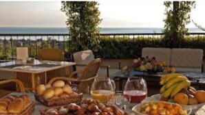 A breakfast buffet with bread, pastries, fruits, and juice on an outdoor table with a view of the sea.