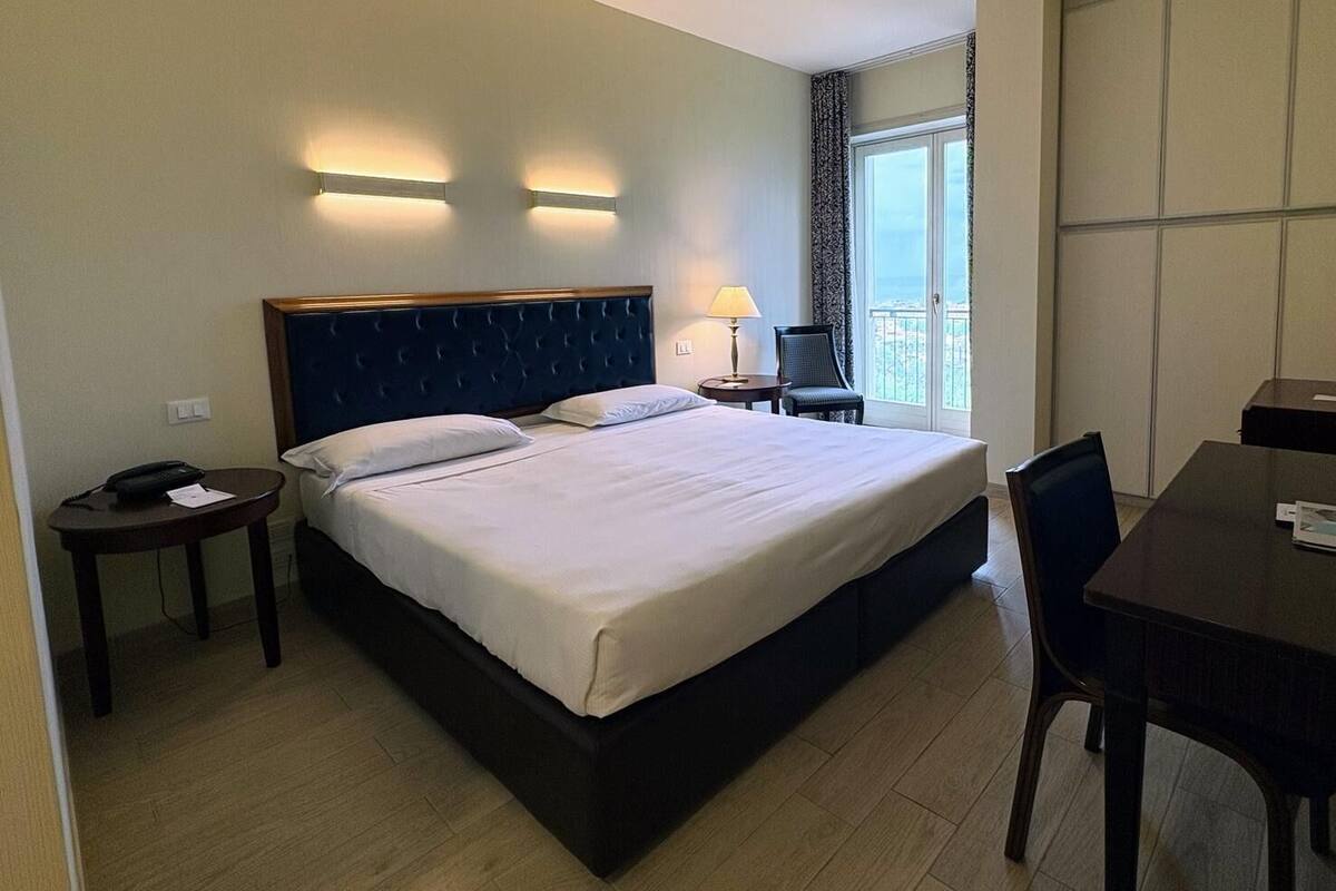 A hotel room with a large bed, a desk, a chair, a lamp, and a view of the city through the window.