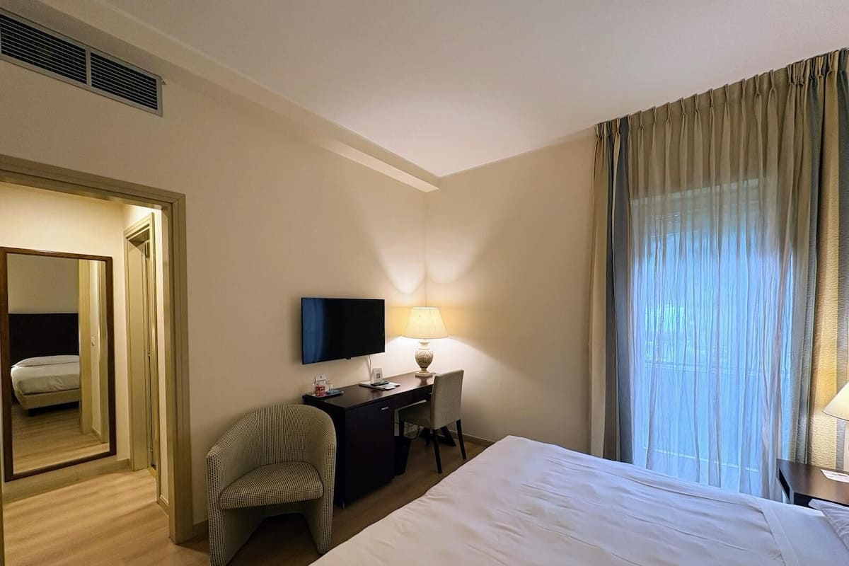 A hotel room with a bed, a desk with a chair, a TV, and a window with curtains.