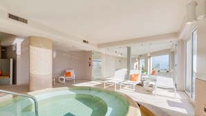 A modern indoor pool area with a glass enclosure, lounge chairs, and a view of the city.