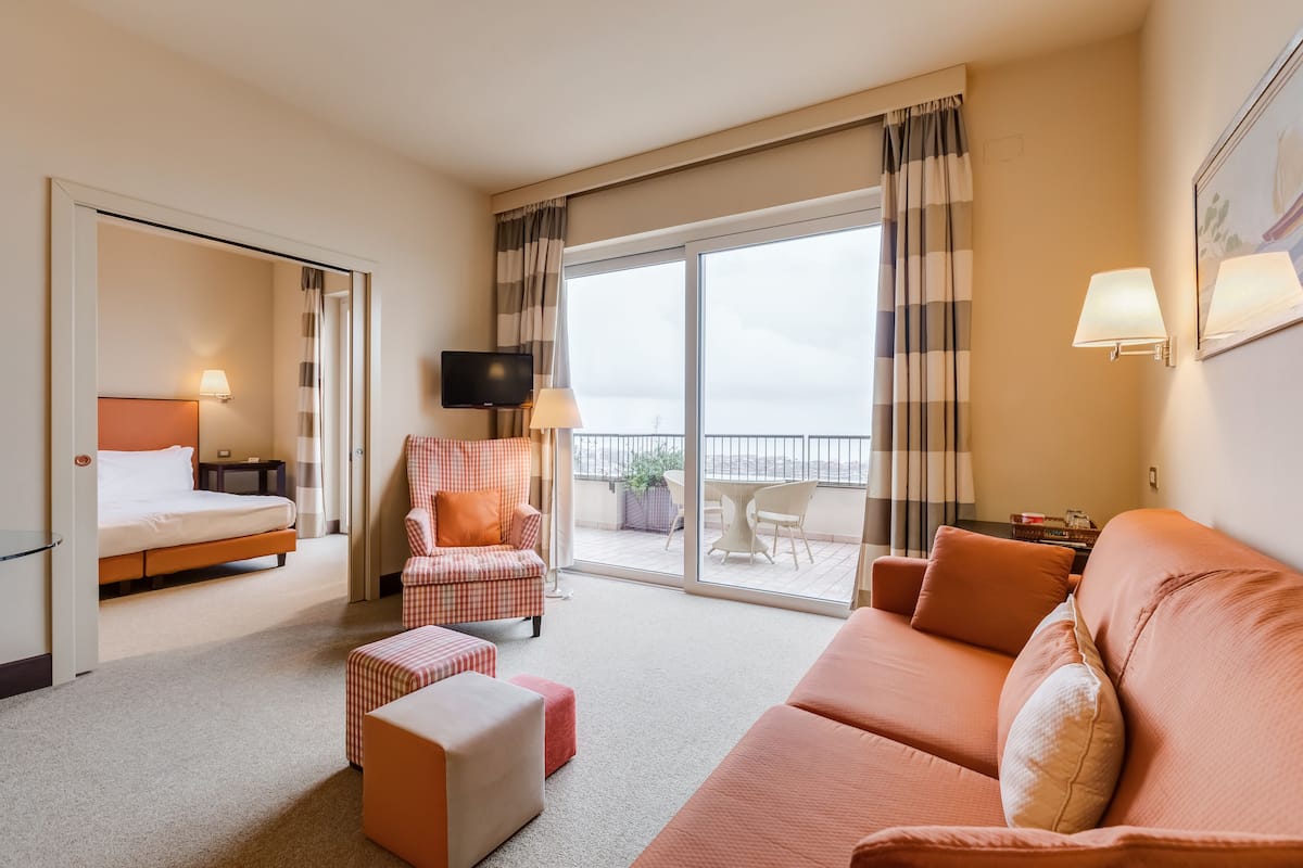 A hotel room with a bed, a sofa, a chair, a TV, and a balcony with a view.