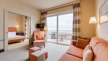 Habitación de hotel con cama, sofá, sillón, televisor y balcón con vistas.