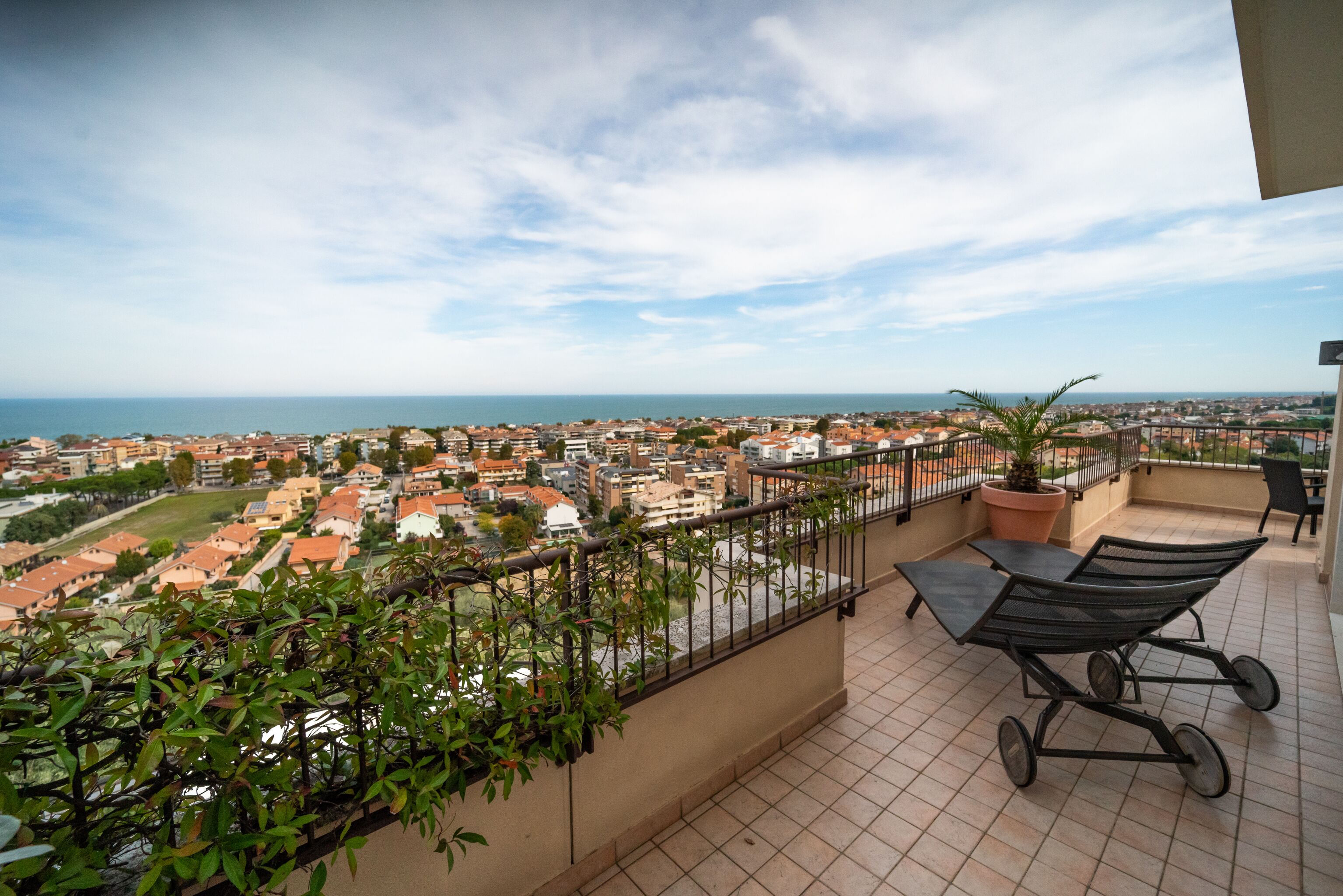 A balcony with a view of the sea and town, a potted plant, and a metal chair.