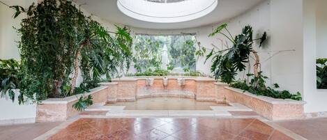 A spacious indoor atrium with a central fountain, surrounded by lush greenery and a high ceiling with a skylight.
