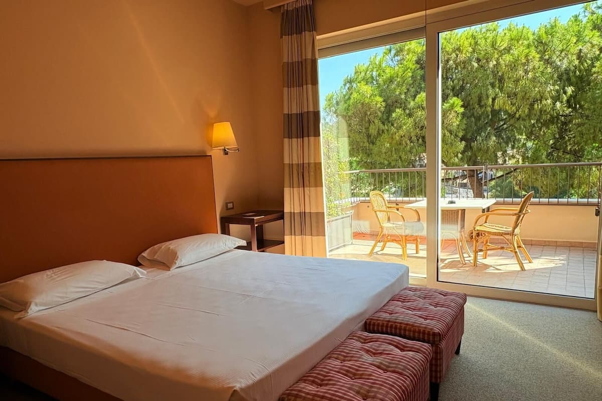 A hotel room with a large bed, a small table, a chair, and a balcony with a table and chairs.