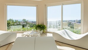 A modern living room with large windows, a white sofa, a white coffee table, and a potted plant.