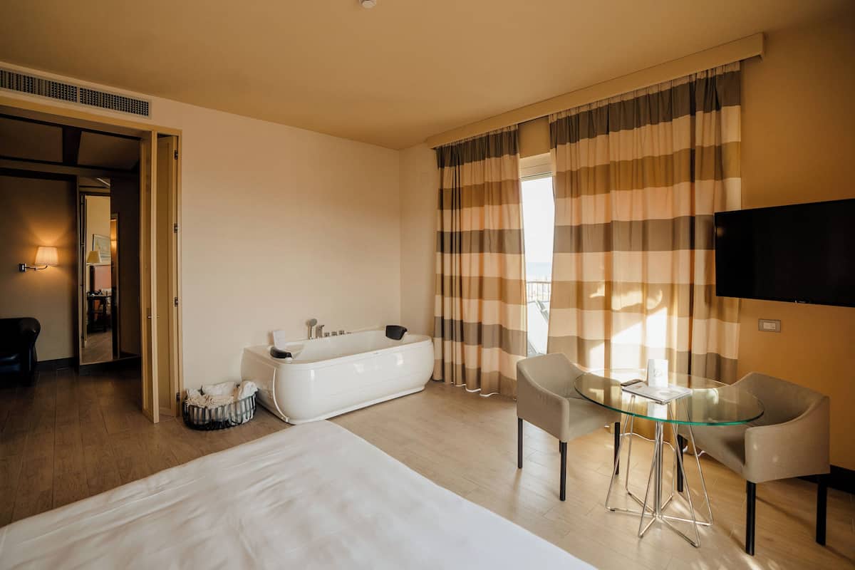 A hotel room with a large bed, a glass table with two chairs, a bathtub, and a television on the wall.