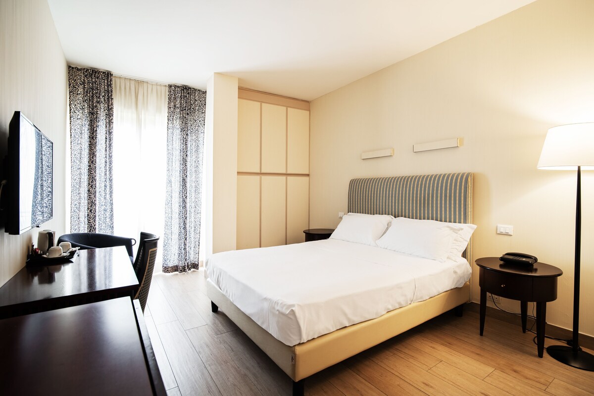 A hotel room with a bed, a desk, a chair, a lamp, and curtains.