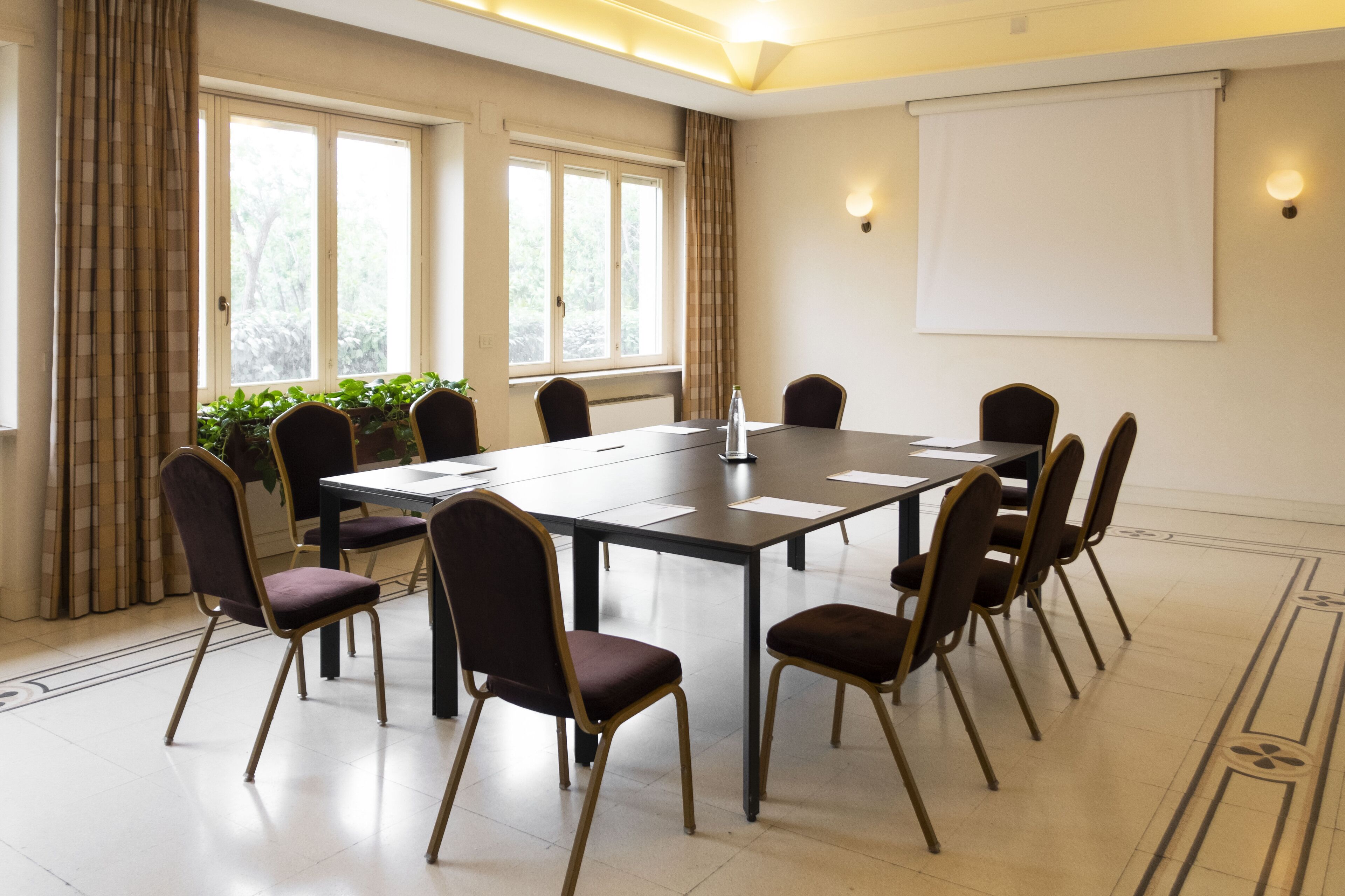 Un salón de conferencias con una mesa larga, sillas, una pantalla de proyector y ventanas con cortinas.