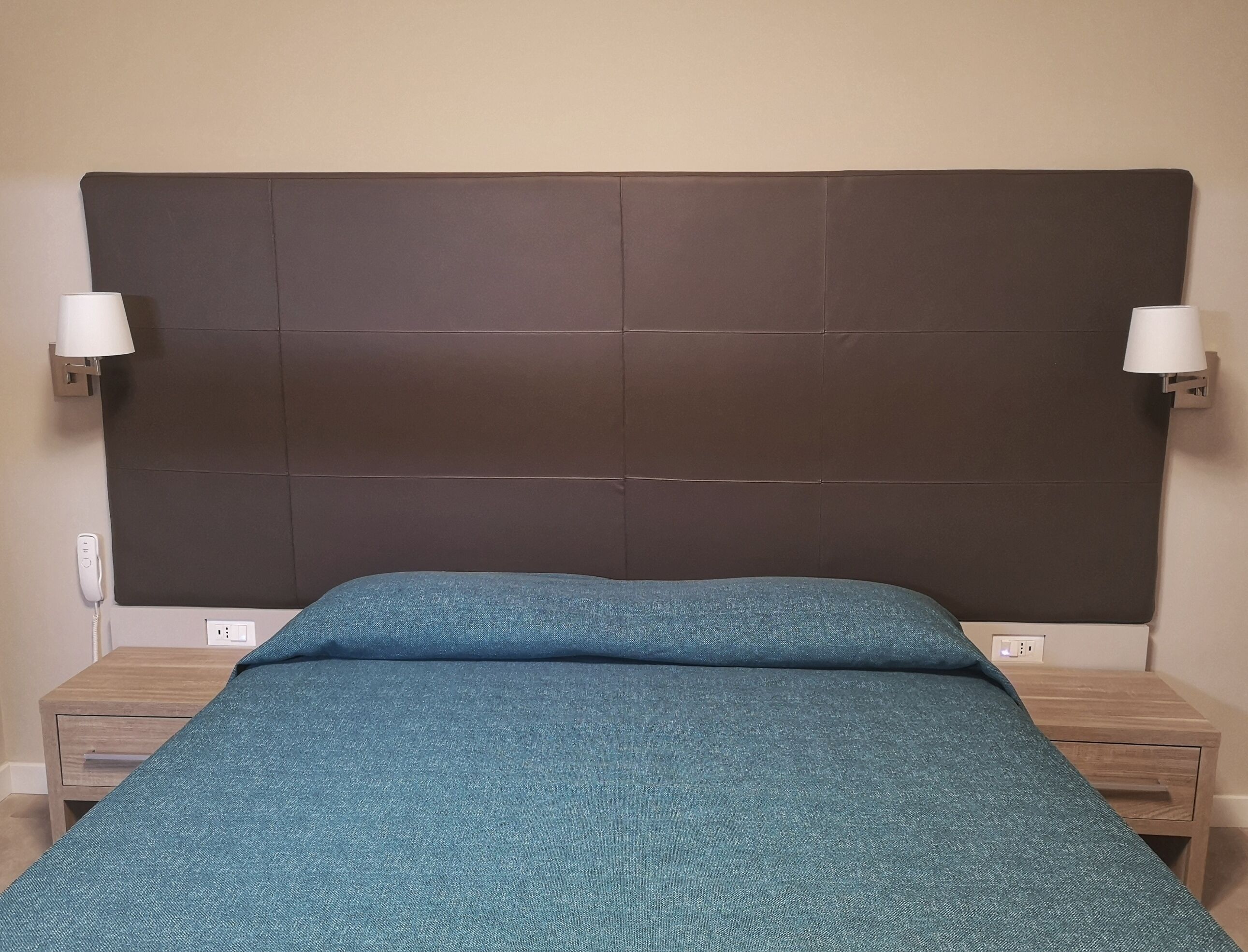 Een slaapkamer met een donker hoofdbord, een blauwgroene bedsprei, twee nachtkastjes en wandlampen.