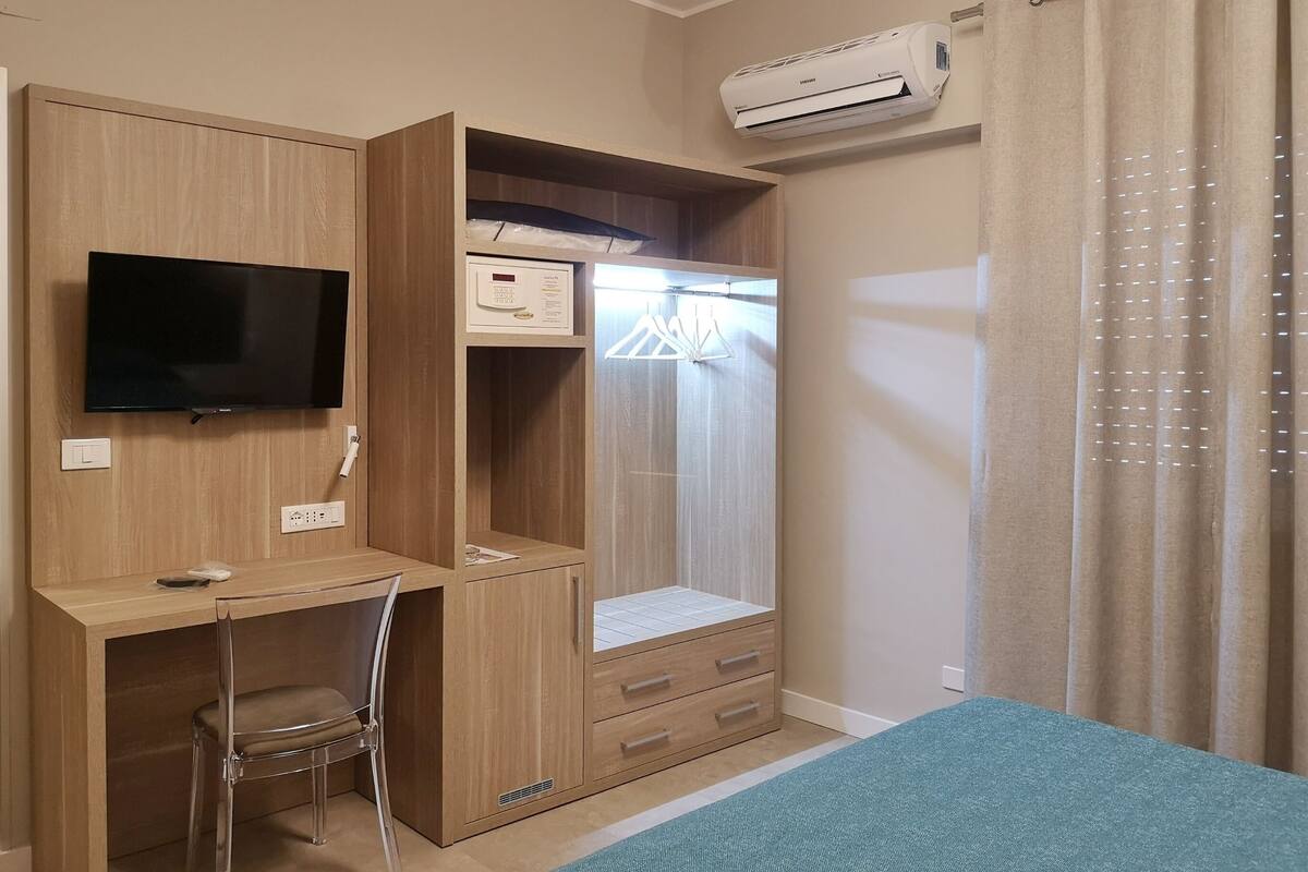 A bedroom with a bed, a desk with a chair, a wardrobe, and an air conditioning unit.