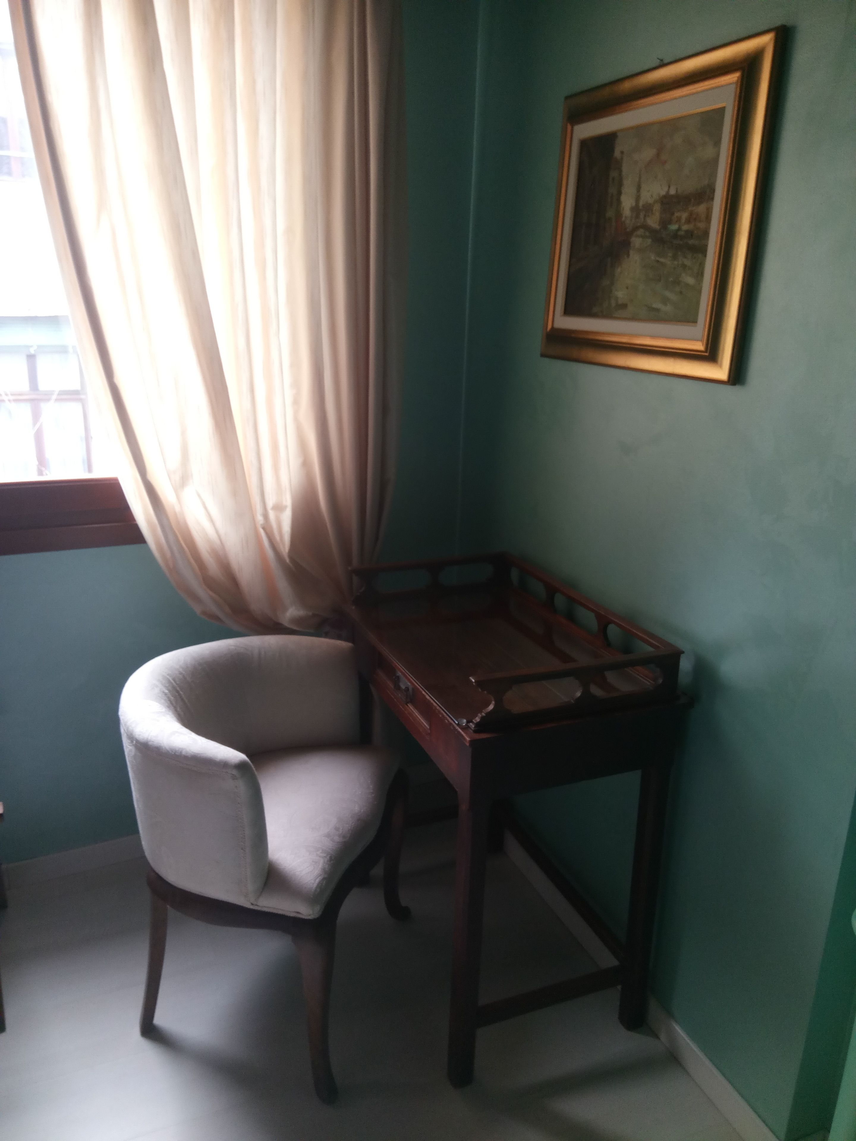 Habitació d'angle amb una cadira, una taula de fusta, un quadre a la paret i una finestra amb cortines.