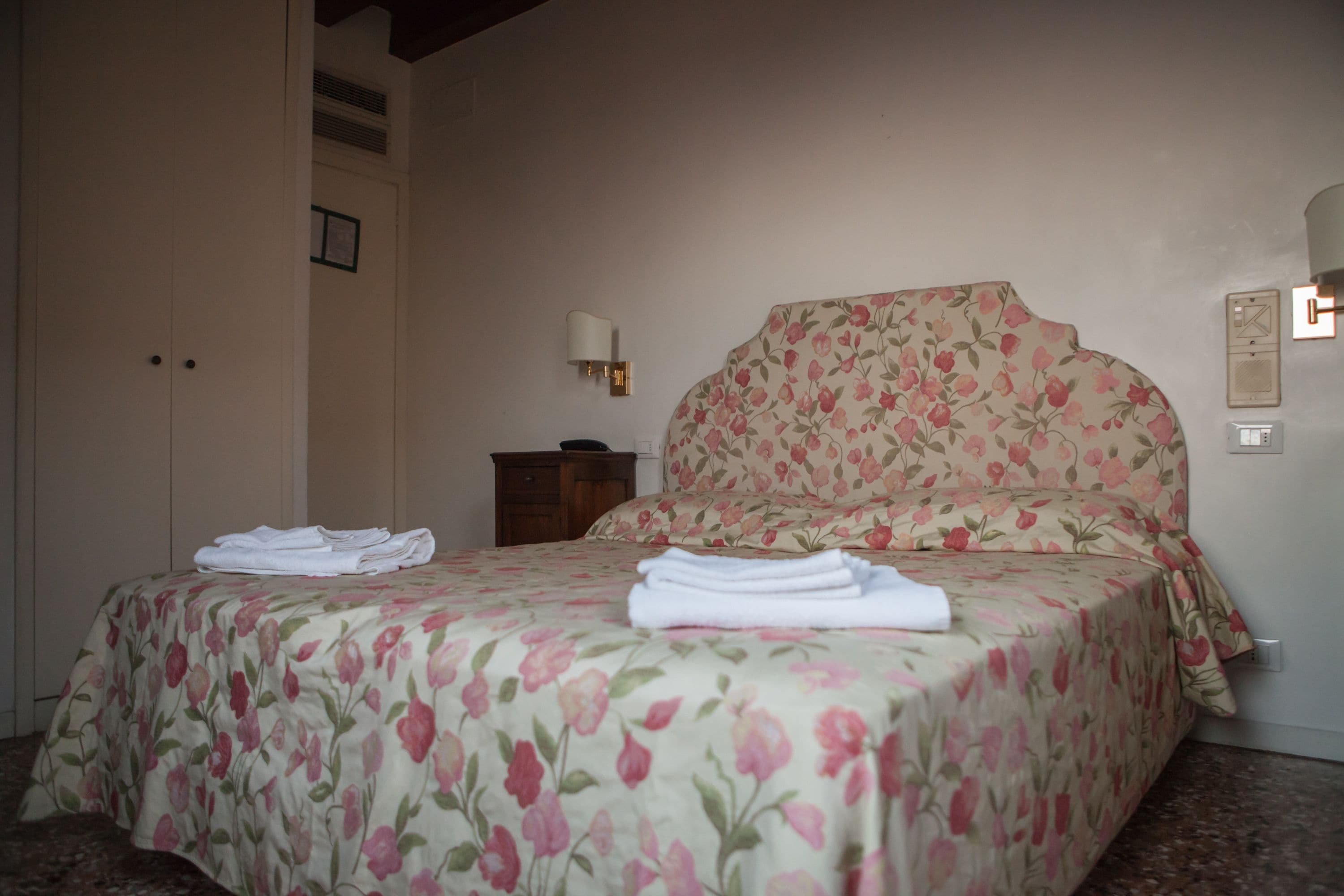 Ein Schlafzimmer mit floralem Überwurf, einem ordentlich gefalteten weißen Handtuch auf dem Bett, einem Nachttisch mit Lampe und einem an der Wand befestigten Thermostat.