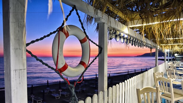 A beachside restaurant with a view of the ocean at sunset, featuring tables and chairs along the railing.