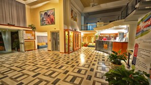 A hotel lobby with a patterned floor, a reception desk, a staircase, and a large painting on the wall.