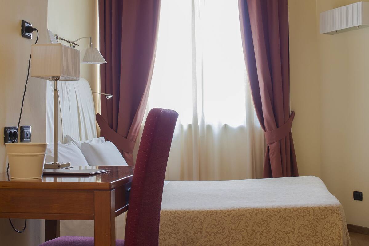Une chambre d’hôtel avec un lit, un bureau avec une lampe, une chaise et une fenêtre avec des rideaux.