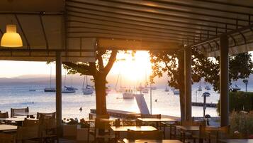 Zona exterior con mesas y sillas, con vista a un puerto deportivo con barcos y un atardecer.