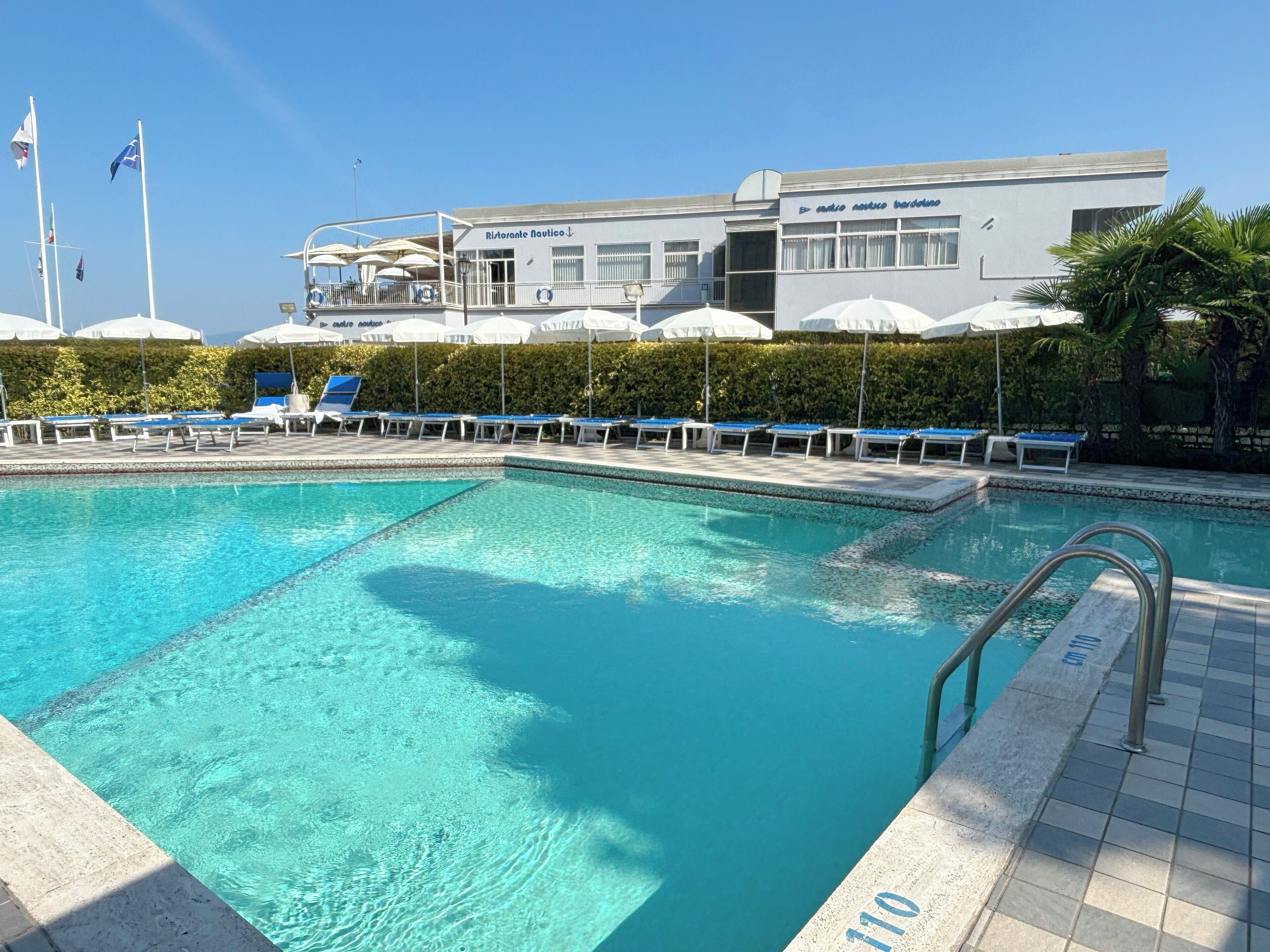 A swimming pool with lounge chairs and umbrellas, a building with a sign reading 'Ristorante Nautico,' and flags in the background.