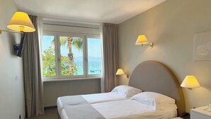 Un cuarto de hotel con cama, dos lámparas de noche, una mesita de noche, televisor y ventana con vista al mar y palmeras.