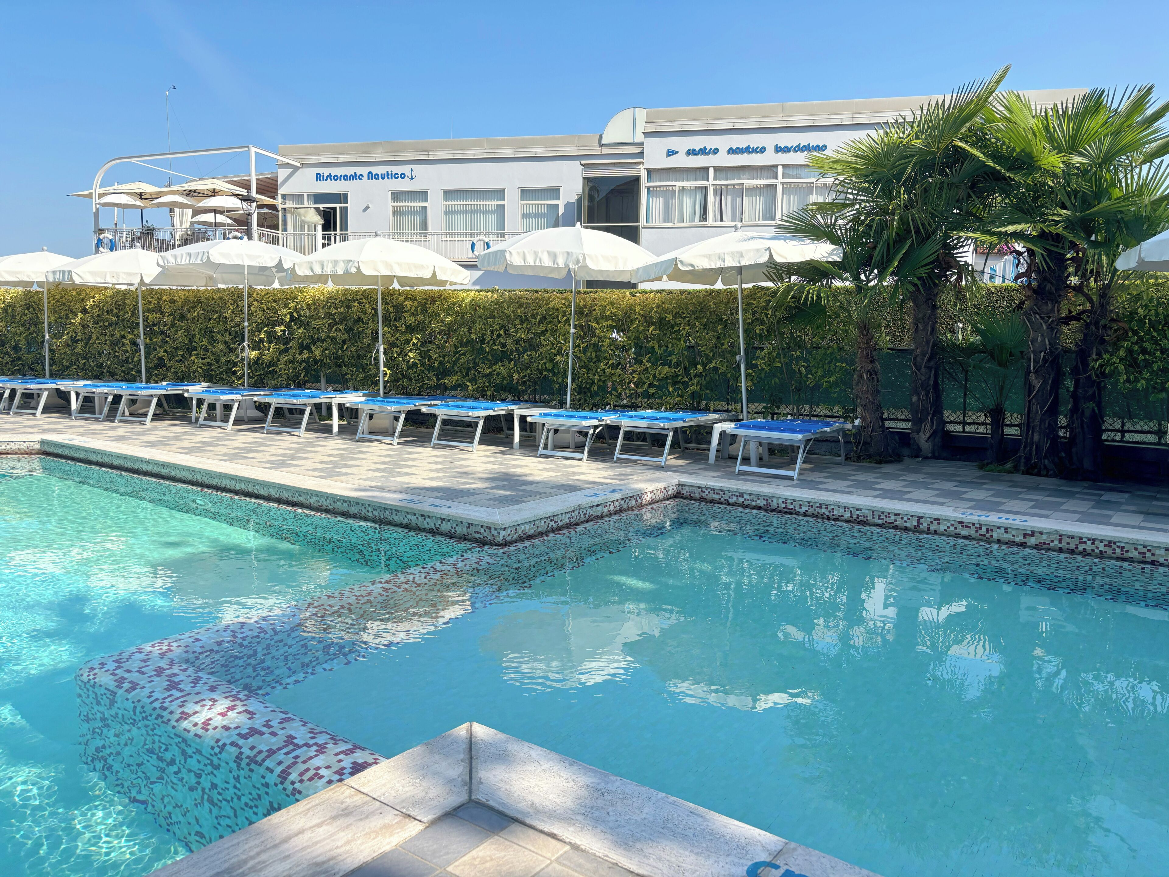 Una piscina con lettini e ombrelloni, un edificio a tema nautico e un siepe curata.