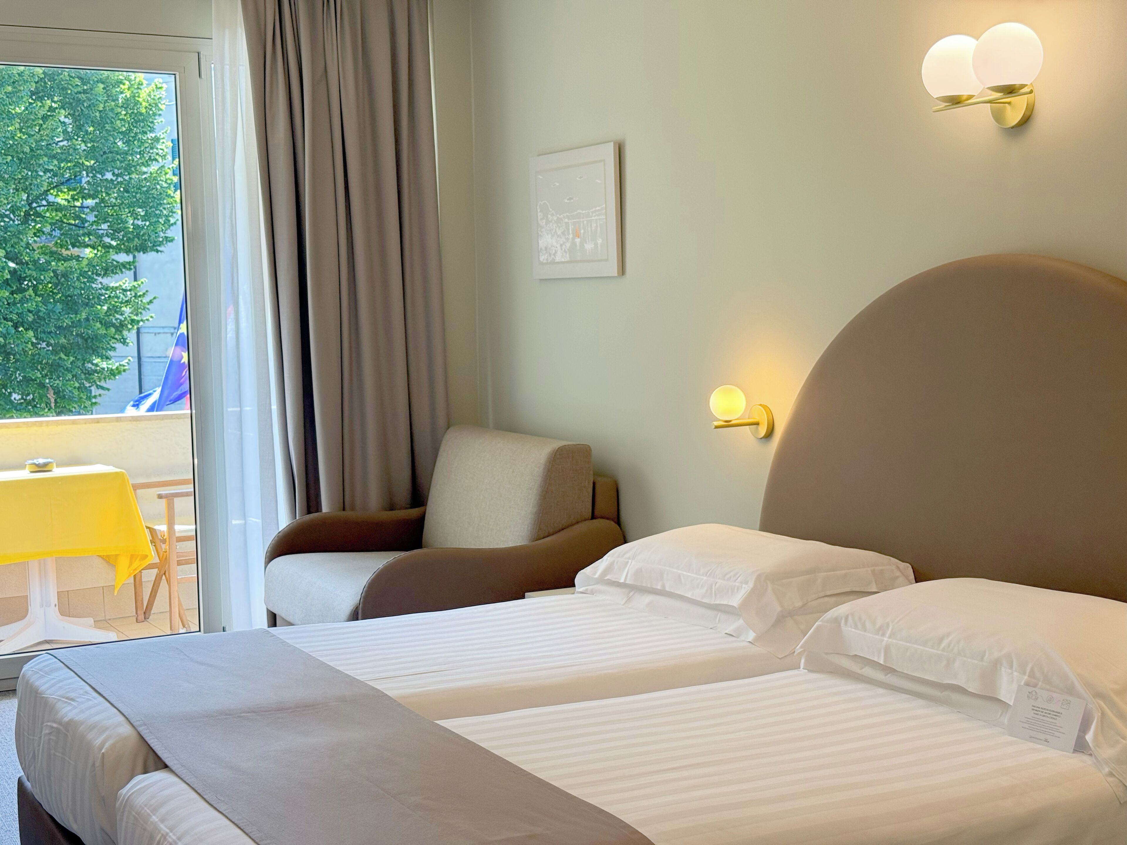 A hotel room with two beds, a chair, a table, and a view of the outdoors.