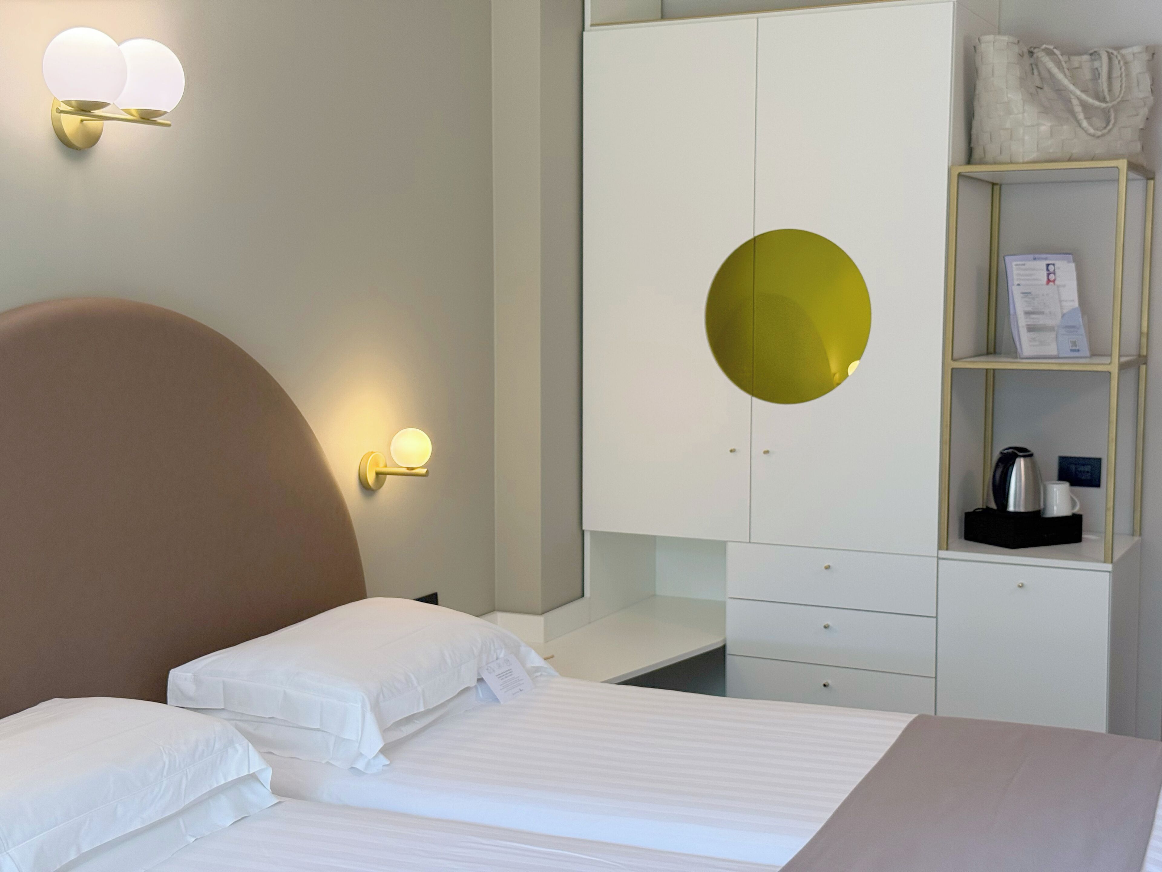 A hotel room with a bed, a wardrobe, a shelf with a kettle and a mug, and wall sconces.