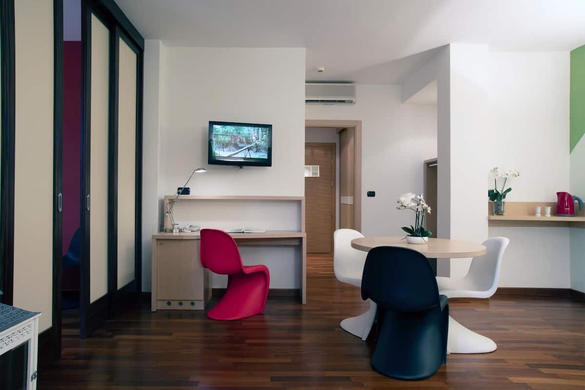 Una habitación de hotel moderna con suelo de madera, un escritorio con computadora, un televisor en la pared y un área de comedor con sillas.