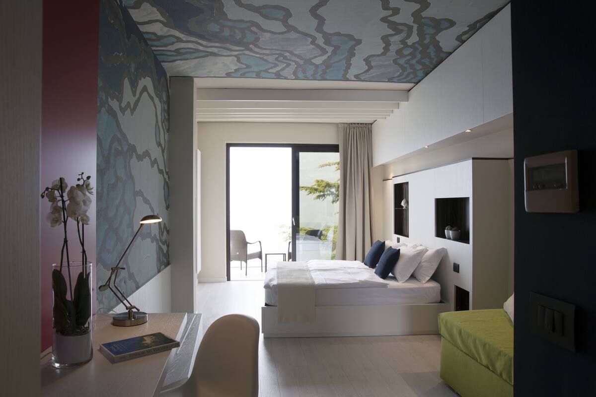 Un dormitorio moderno con cama, escritorio, silla y un taburete verde. Se ve un ventanal con vistas al exterior.