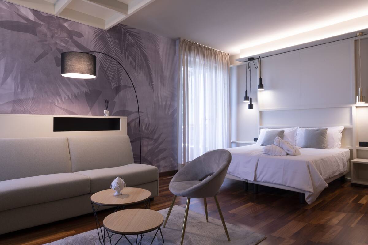 Una habitación de hotel moderna con sofá, una silla, una mesa redonda, una cama con almohadas y una mesita de noche.