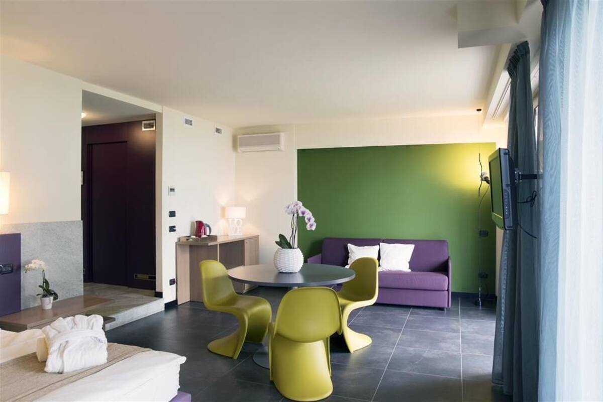 Una habitación de hotel moderna con zona de comedor, un sofá púrpura, una pared de acento verde y un televisor.