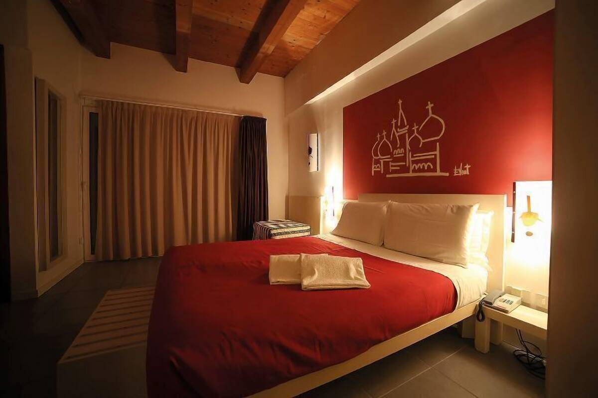 Een hotelkamer met een groot bed, een rode muur met een wit grafisch element, een nachtkastje met een lamp en een raam met gordijnen.