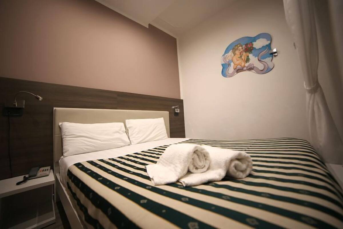 Een hotelkamer met een bed, nachtkastje, wandklok en een decoratieve muurschildering van een engel.