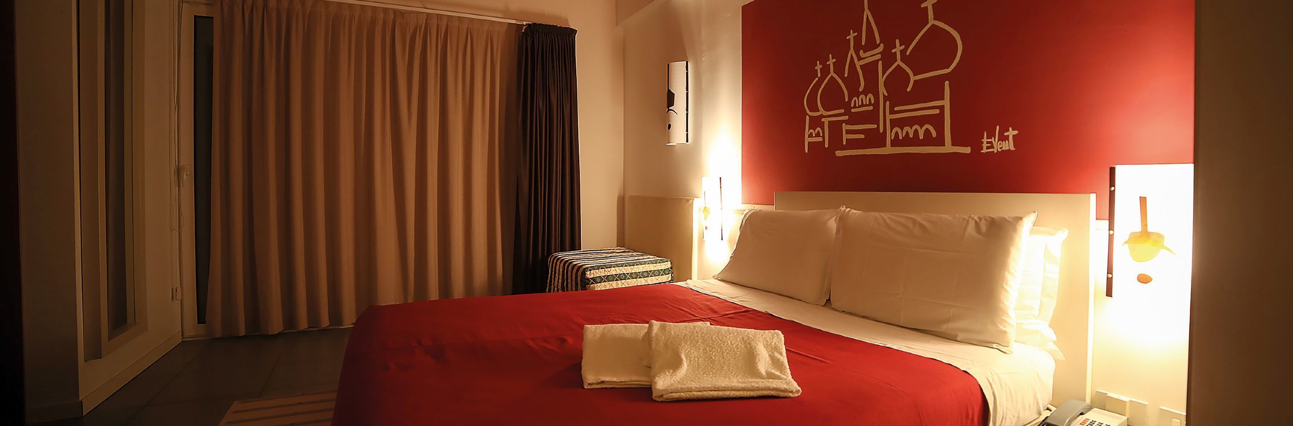 Une chambre d’hôtel avec une couvre-lit rouge, une petite table de chevet sur laquelle repose un plateau décoré, et un mur orné d’un simple dessin linéaire.