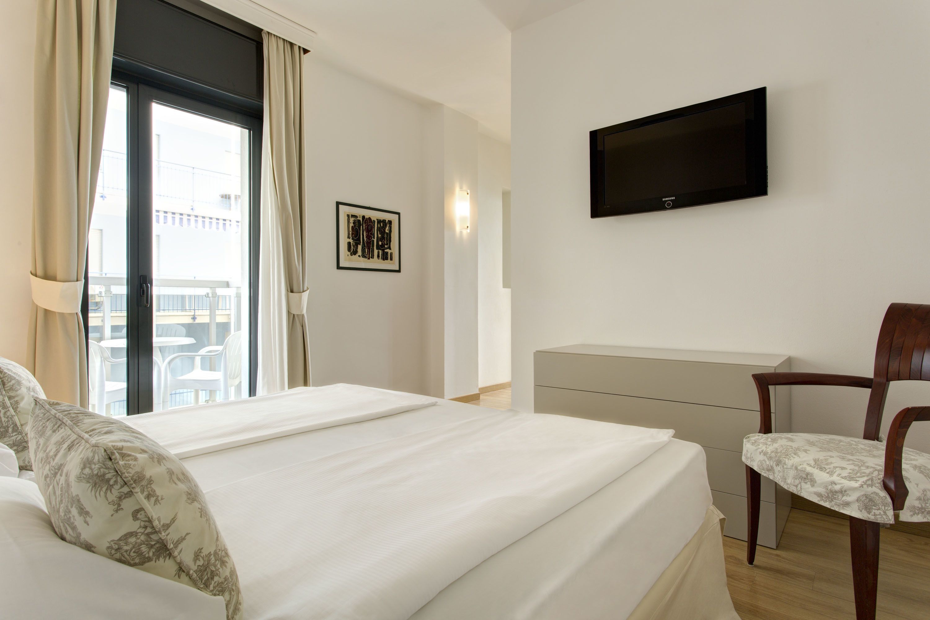 A hotel room with a bed, a desk, a chair, a TV on the wall, and a window with curtains.
