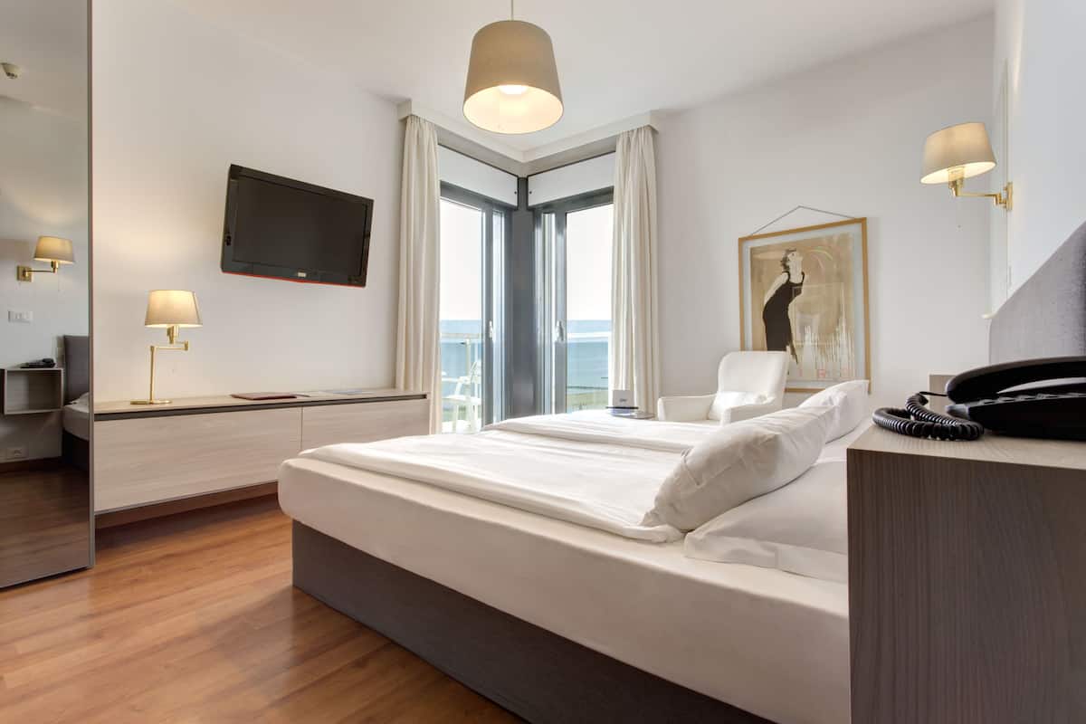 A modern hotel room with a large bed, a desk with a flat-screen TV, a chair, and a view of the sea through a large window.