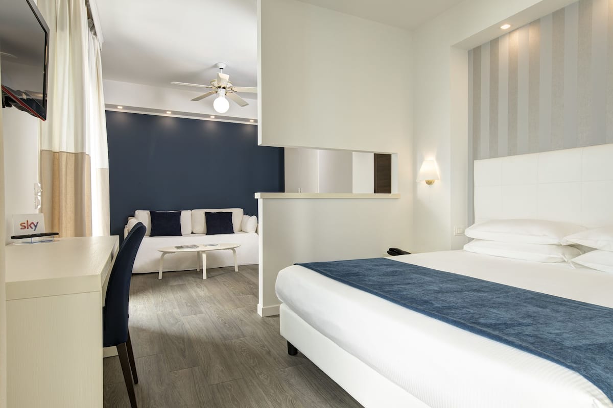 A modern hotel room with a bed, a desk, a chair, a small table, a ceiling fan, and a wall-mounted TV.