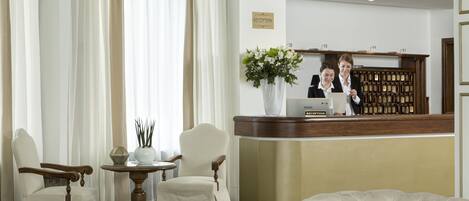 A reception area with a reception desk, two chairs, a small table with a vase of flowers, and a large window with curtains.