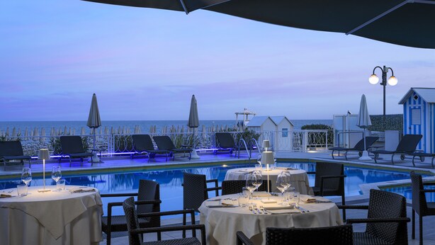 A poolside dining area with tables set for two, lounge chairs, and umbrellas. - Hotel Byron Bellavista (Jesolo)