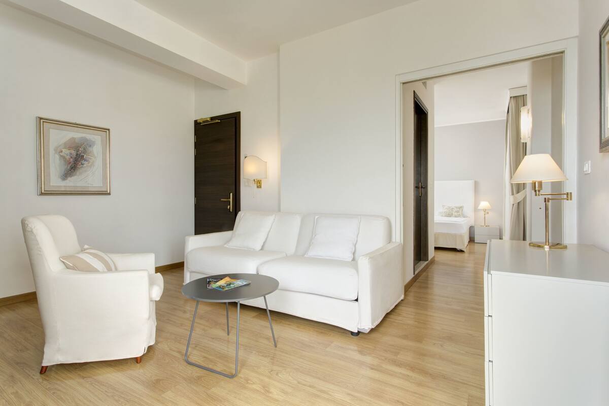 A modern living room with a white sofa, a white armchair, and a small round table. There is a door leading to another room and a lamp on the right side.